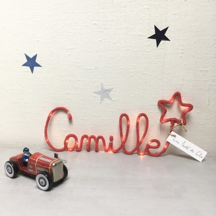 Prénom Camille réalisé en tricotin lumineux en fil de coton de couleur rouge présenté sur un meuble accompagné d'une vieille voiture de course. trois étoile sont sur le fond du mur