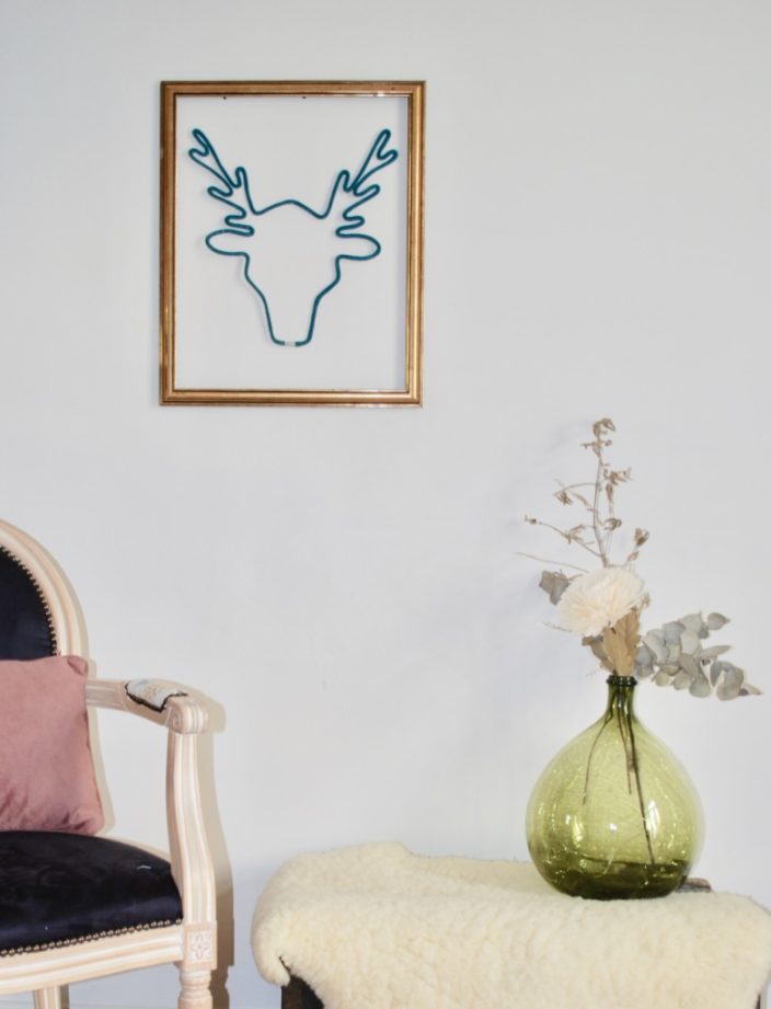 trophée grand cerf en tricotin couleur pin dans un cadre en bois, accroché au mur d'un salon blanc. salon décoré d'une dame jeanne fleurie posée sur un peau de mouton. ert meublé d'une chaise style louis XVI bleu marine